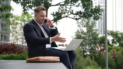 Caucasian businessman sitting at eco park while talking by using phone and working on laptop....