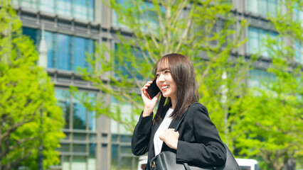 新緑の街で歩きながらスマホで通話する女性ビジネスパーソン