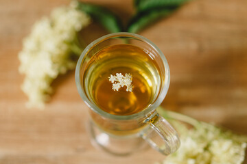 A clear glass mug filled with elderflower tea is placed on a rustic wooden table. Fresh elderflowers and a wooden cutting board are nearby, creating a cozy and natural atmosphere.