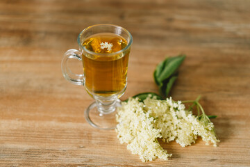 A clear glass mug filled with elderflower tea is placed on a rustic wooden table. Fresh elderflowers and a wooden cutting board are nearby, creating a cozy and natural atmosphere.