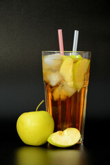 A faceted tall glass of apple juice with ice on a black background, next to ripe slices of a green...