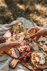Close up of two people's hands toasting and clinking white wine glasses over charcuterie board spread on blanket at picnic in the park, Generative AI