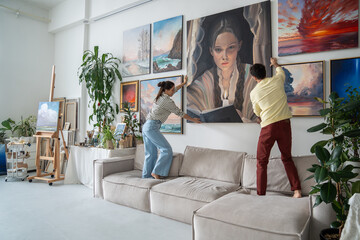 Married talented couple hangs trendy art canvas on wall in spacious bright living room gallery....