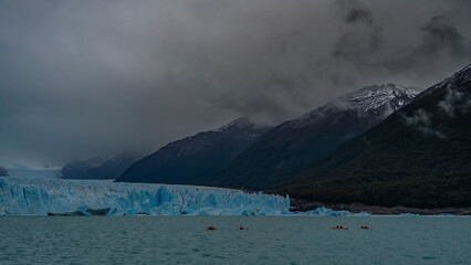 A wall of blue ice in a turquoise glacial lake. The Perito Moreno glacier stretches to the horizon between the mountains. Canoes with tourists are sailing. Cloudy. Fog. El Calafate. Argentina.  