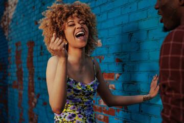 A joyful woman with curly hair laughing and dancing in front of a blue brick wall, interacting...