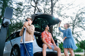 Little boy holding EV charger and point at camera with his family sitting on the trunk in...
