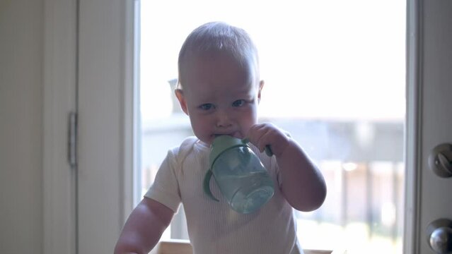 An adorable toddler stands at learning tower, drinking independently from a sippy cup, captured in a moment that highlights early self-feeding skills and joy of little achievements in a home setting