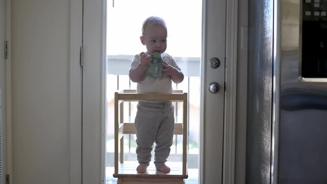 toddler stands in a wooden learning tower by open doorway happily sipping from green sippy cup natural light illuminates the scene, capturing a moment of independence and early childhood development.