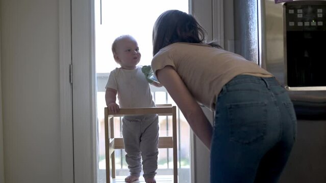 This tender scene captures a toddler in a learning tower offering a sippy cup to his mother, highlighting a moment of sharing and connection in a sunlit kitchen, symbolizing early social development.