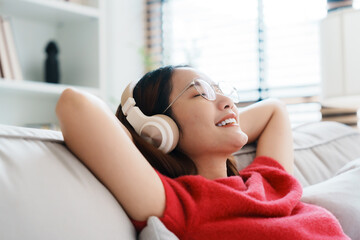 Relaxed young Asian woman enjoying rest on comfortable sofa, calm attractive girl relaxing on couch, breathing fresh air with eyes closed, wearing eyeglasses and headset, meditating at home