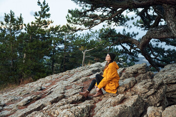 Serene woman in a vibrant yellow coat gazes at the majestic ocean and lush trees from mountain top