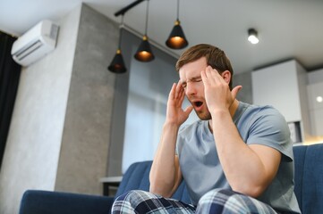 Unpleasant pain. Sad unhappy handsome man sitting on the sofa and holding his forehead while having...