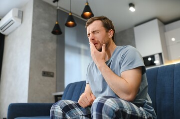 Toothache. Unhappy upset caucasian sad man sits on the couch at home, holds his hand near his...