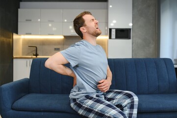 People, Healthcare and Treatment. Portrait of sick man suffering from acute side back pain, sitting on couch at home, person touching lower back aching area with hand. Health Problem Concept
