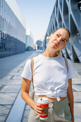 A 30-year-old young woman stands at the business center of the city with a cup of coffee and looks up thoughtfully