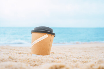 A paper cup with coffee on the sand overlooking the sea. The concept of morning and breakfast with cappuccino on vacation by the sea