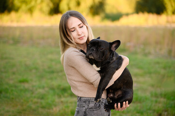 While walking in park, young dog owner holds her beloved pet, French bulldog, in her arms and looks at him lovingly. Concept of people, animals, friendship and loyalty, caring for dog.