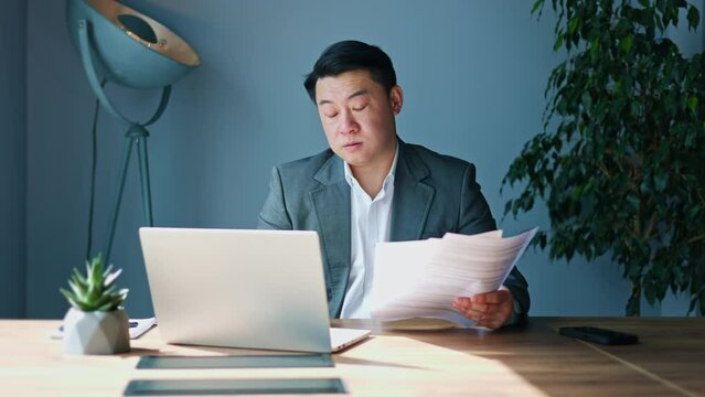 Sleepy young multitask male looking at display of portable laptop, reading documents and cheking personal smartphone in designer cabinet with blue walls. Concept of technology and occupation.