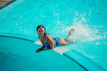A woman is in the water, smiling and enjoying herself. The water is blue and the pool is surrounded...