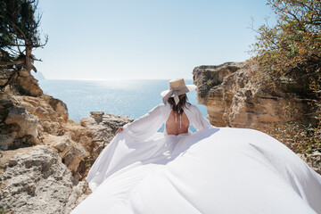 A woman in a white dress is standing on a rocky cliff overlooking the ocean. She is wearing a straw...