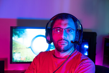 Handsome young man in casual clothes and headset sitting at a table in a room with purple lights and playing video games on the computer with a smile on his face looking at the screen