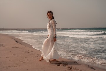 Model in boho style in a white long dress and silver jewelry on the beach. Her hair is braided, and there are many bracelets on her arms.