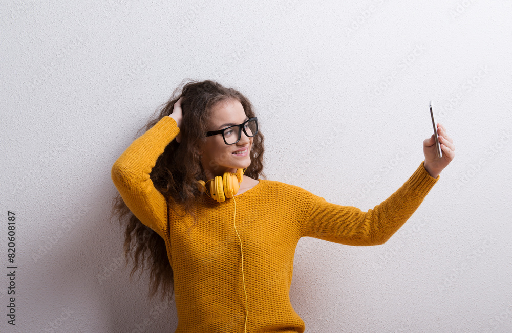 Wall mural Portrait of a gorgeous teenage girl with curly hair and headphones,taking selfie. Studio shot, white background with copy space.