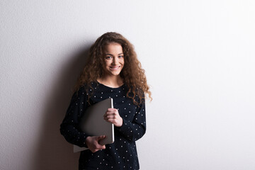 Portrait of a gorgeous teenage girl with curly hair, holding notebook, leaning against wall. Studio...