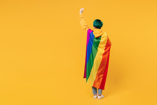 Full body back rear view young lesbian woman with dyed green hair wrapped in rainbow flag raising up hands clench fist isolated on plain yellow orange wall background LGBT diversity lifestyle concept