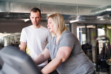Overweight woman exercising on treadmill in gym. Personal trainer helping her.