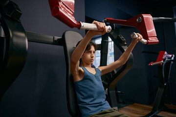 Active athletic preteen boy training arms muscles at gym