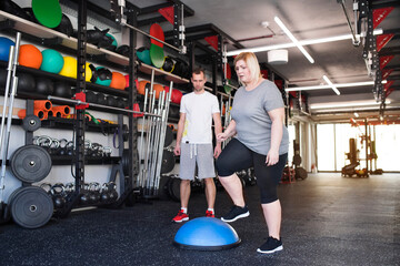 Overweight woman exercising on balance ball in gym. Personal trainer couching her and helping her.