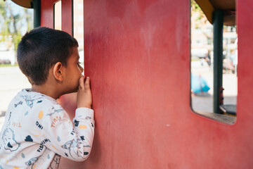 Curious autistic child peeking through window at playground. Young boy looks through red window...