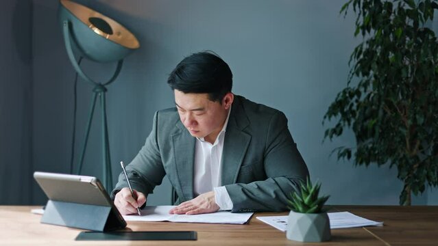 Reflective chinese executive carefully reviewing documents at assigned workspace in office environment. Diligent male gesturing with creativity and insightful thoughts indoors. Concept of work.