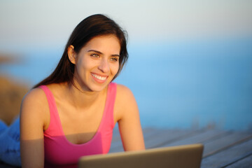 Happy woman with a laptop looking away at sunset on the beach