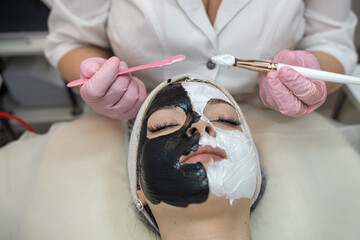 Beautiful young woman receive white and black facial mask on half face lying on spa