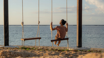 A woman is seen from behind, swinging over the ocean as the sunsets on the beach.