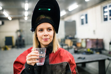 Beautiful blonde woman works as a welder in workshop, drinking water from glass. Wearing protective...