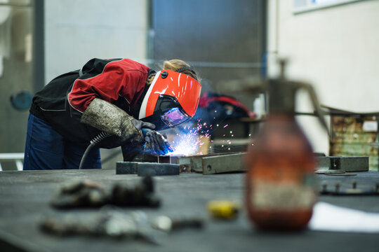 Beautiful blonde woman works as a welder in workshop, operating welding machine, wearing protective clothing and a welding mask.