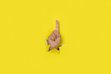 Hand making a sign of number one, coming out of the hole in a torn yellow paper background.