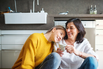 Mother and mature daughter in kitchen, sitting on floor and talking. Closeness of mom and her adult...