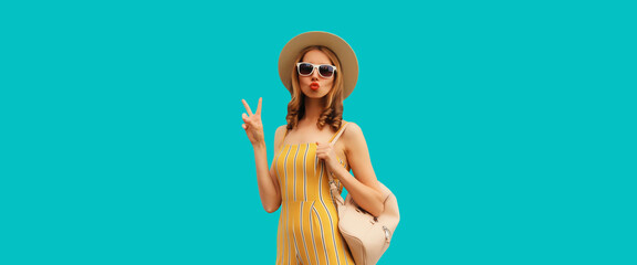 Beautiful blonde young woman blowing a kiss posing wearing summer hat, backpack on blue background