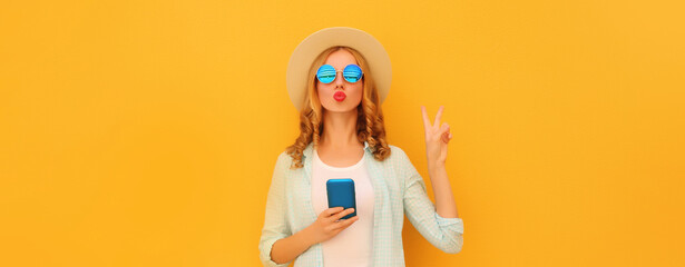 Stylish woman posing blowing a kiss with smartphone wearing summer hat posing on yellow background