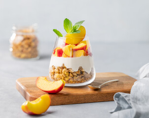 Greek yogurt parfait with peach and granola in a glass on a wooden board on a light background with fresh fruits and napkin.