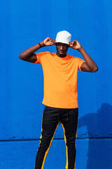 Young African American male in bright yellow t shirt and sportive pants adjusting white cap while standing against blue wall
