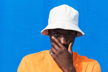 Young African American male in bright orange t-shirt and white cap touching face with hand while standing against blue wall