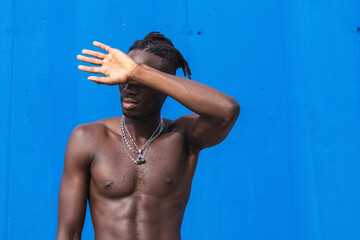 Confident black man standing shirtless over isolated blue background covering his face with hand