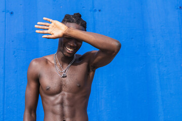 Black man standing shirtless over isolated blue background smiling to the camera and covering his...