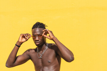 Serious young shirtless African American male in stylish sunglasses and with metal necklaces posing...