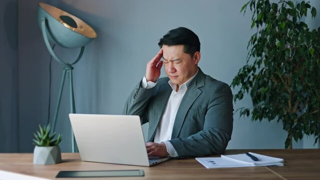 Troubled japanese man massaging temples while brow furrowed with throbbing pain of headache in bustling office. Frustrated young male seeking relief amidst clamor of workspace despite discomfort.
